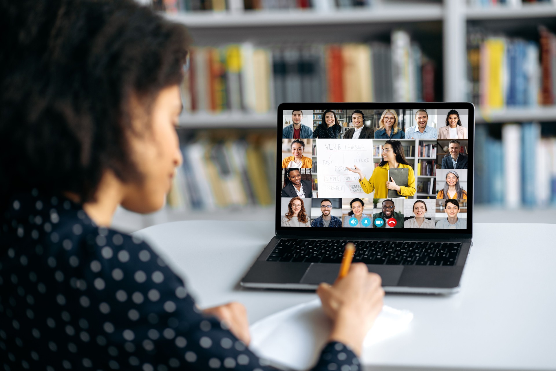 View over shoulder of a mixed race clever female student at a laptop screen with a female teacher and students. Teacher conducts online lecture, female student takes notes. Online training,video call